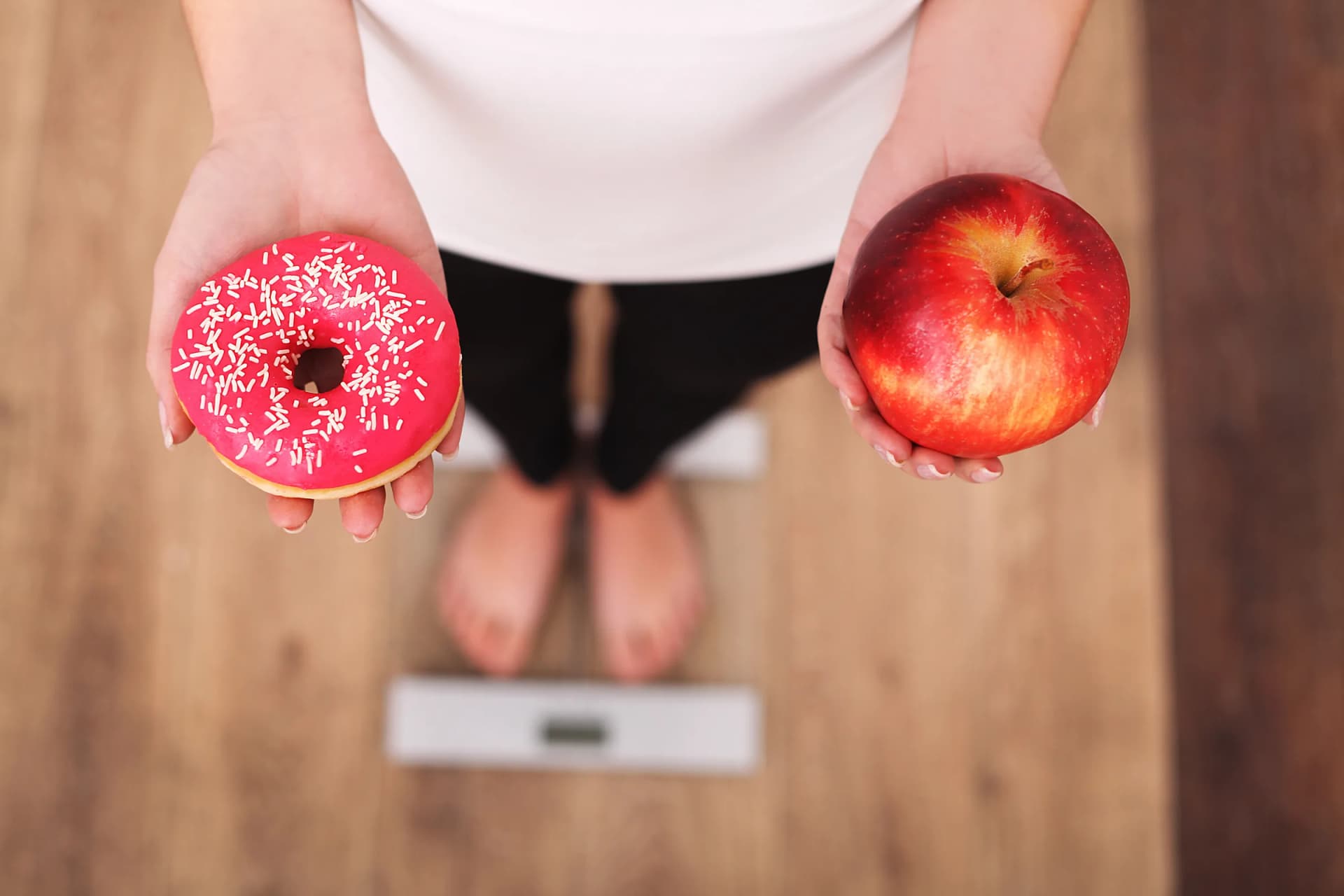 Person on a scale holding a donut in one hand and an apple in the other